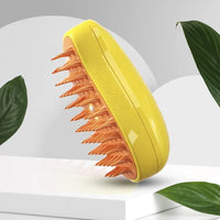 Thumbnail for The KittyNook Cat Co. Electric Cat Grooming Brush with orange bristles is shown on a white stand among green leaves.
