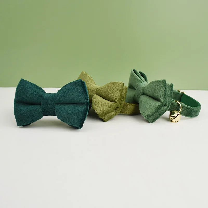 Three green fabric bowties and a KittyNook Cat Co. Velvet Cat Collar with a gold bell on a white and green background.