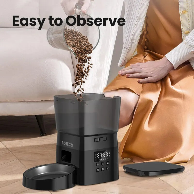 A woman pours food into a black KittyNook ROJECO Automatic Pet Food Dispenser with Smart Control near a sofa.