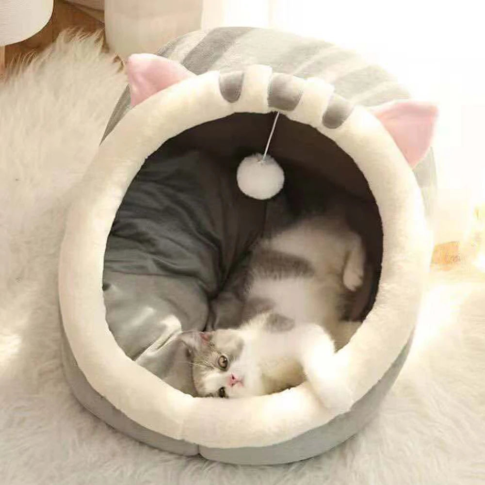 A gray and white cat lounges belly up in the KittyNook Cat Co. Cute Comfortable Cat Nest Bed with pink ears and a toy.