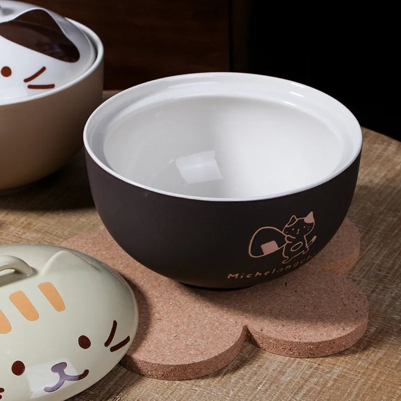 The Cute Cartoon Cat Soup Bowl by KittyNook Cat Co. sits on a cork mat beside a beige lid with a cat face.