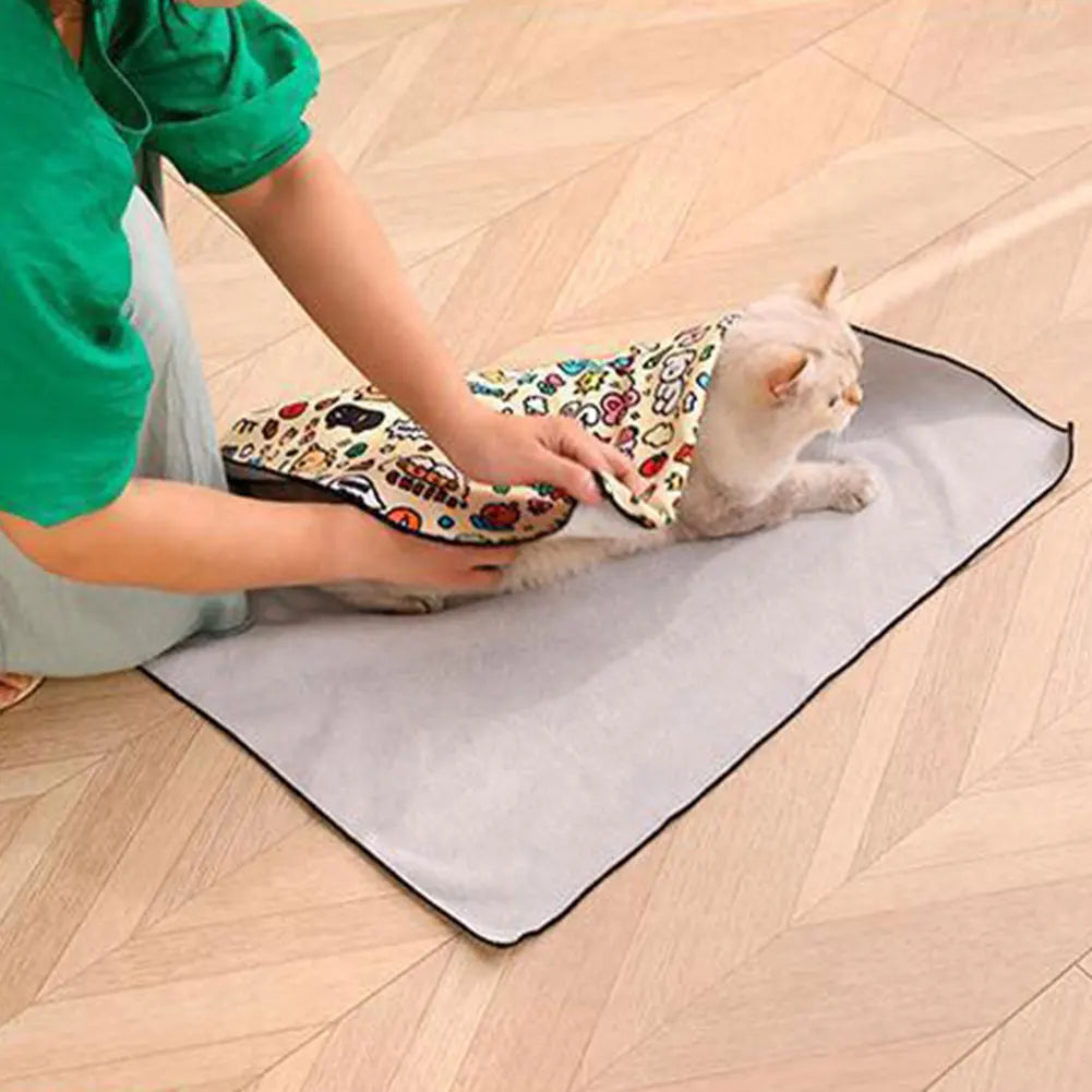 A person wraps a cat in the KittyNook Cat Co. Burrito Swaddle, safely securing it on a mat on a wooden floor.