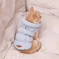 Thumbnail for A ginger cat wears the KittyNook Cat Co. Ultra-Soft Padded Winter Cat Jacket, sitting on a cream cushion, facing away.
