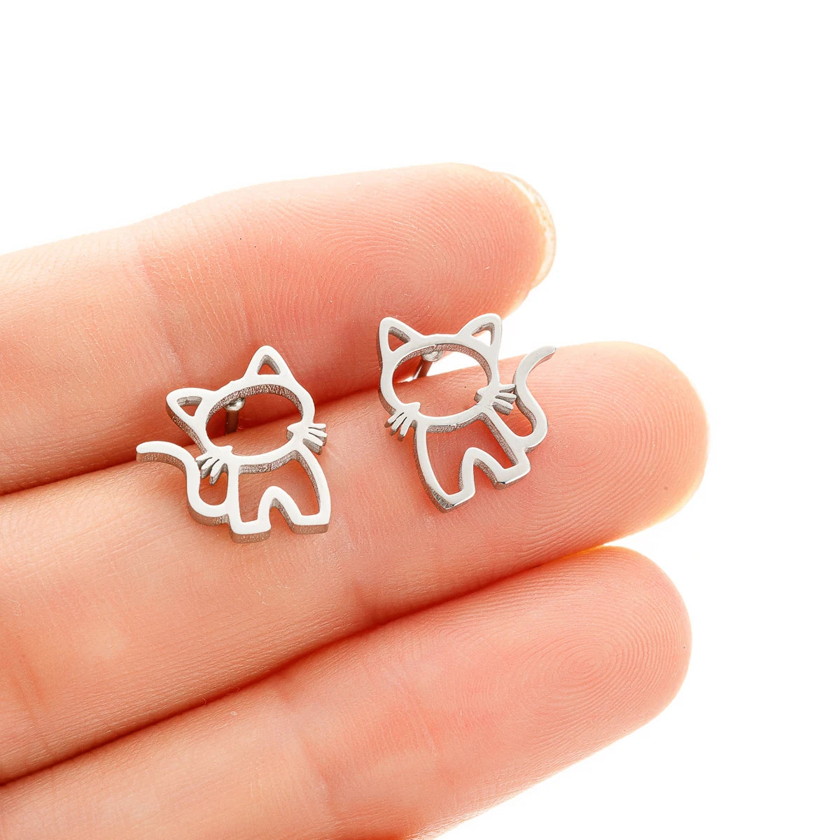 A hand holds two KittyShine Stainless Steel Ear Studs by KittyNook Cat Co., shaped like cute cats, on white background.