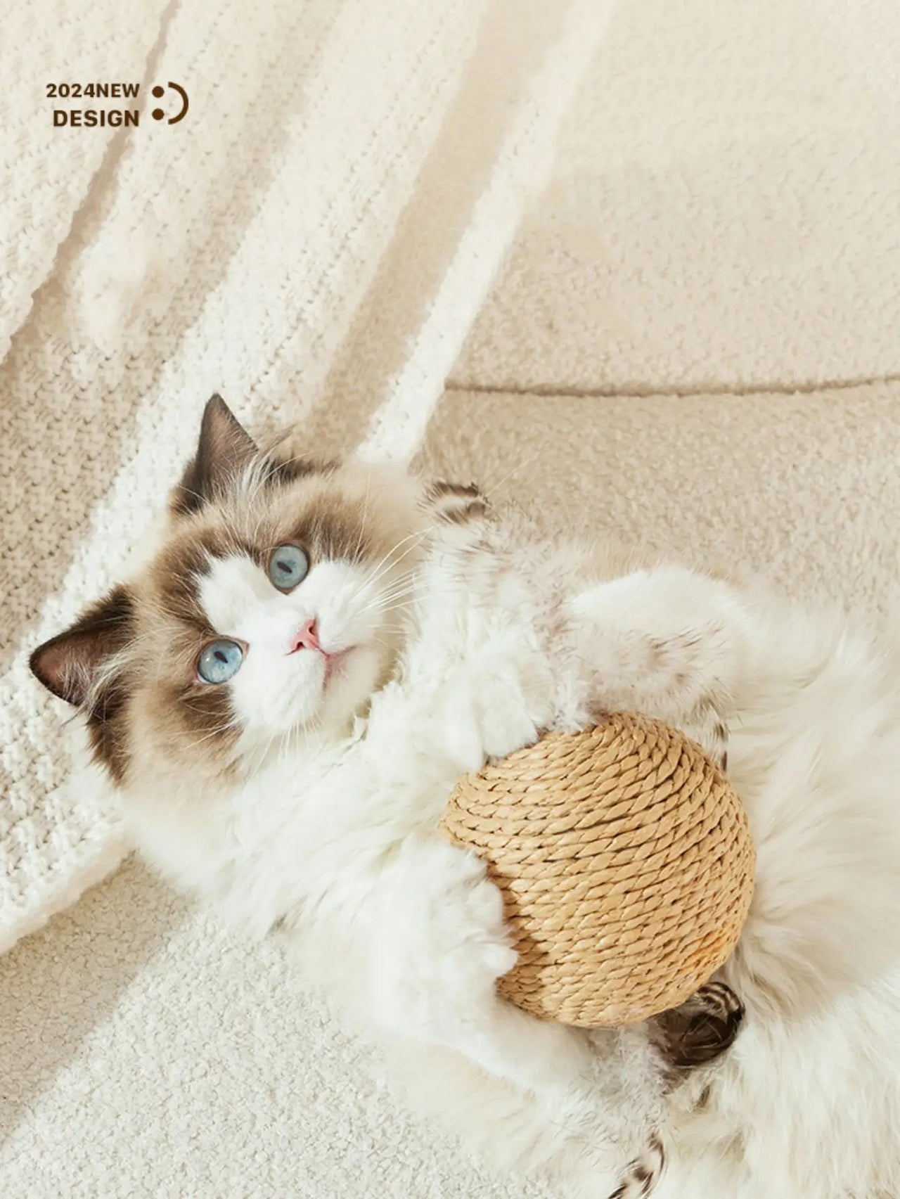 A fluffy white and brown cat with blue eyes lies on a carpet, holding a KittyNook Premium Sisal Kitten Scratching Ball.