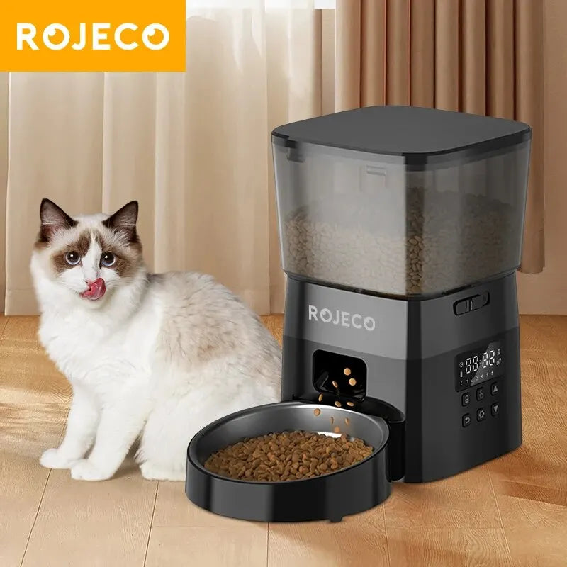 A fluffy cat sits by a ROJECO Automatic Pet Food Dispenser from KittyNook, dispensing food onto the wooden floor.