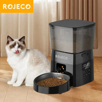 Thumbnail for A fluffy cat sits by a ROJECO Automatic Pet Food Dispenser from KittyNook, dispensing food onto the wooden floor.