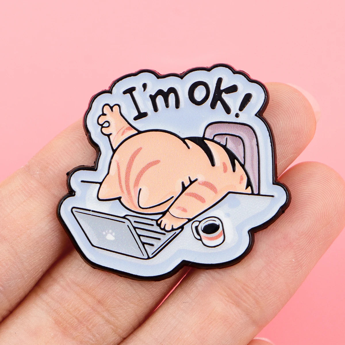 KittyNook Cat Co.'s "Cat at Work" enamel pin features a slumped cat and "I'm OK!" above, shown on a pink background.