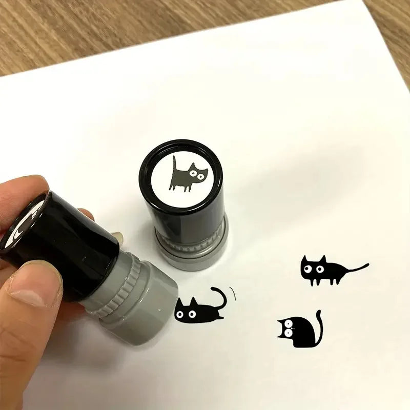 A hand uses the KittyNook Cat Co. Black Cat DIY Journaling Stamp to print a kawaii black cat on white paper.