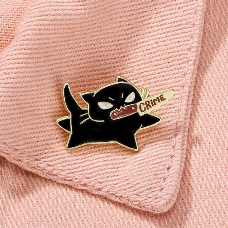 KittyNook Cat Co.'s Cute Black Cat Enamel Brooch shows a cat with a "CRIME" knife on pink textured fabric.