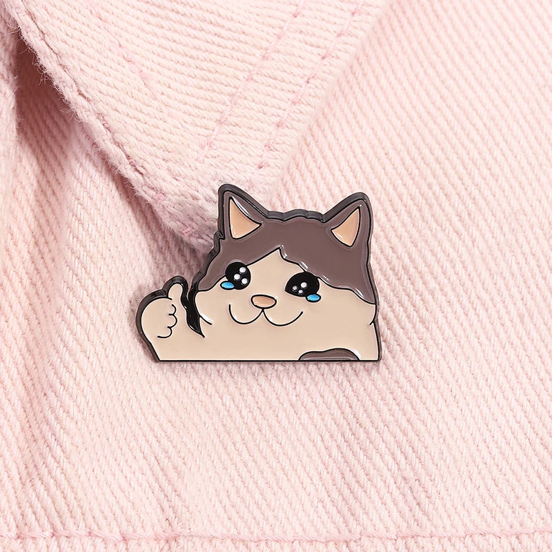 KittyNook Cat Co.'s Kawaii Thumbs Up Crying Cat Enamel Pin on pink fabric—purrfect for cute cat fans!.