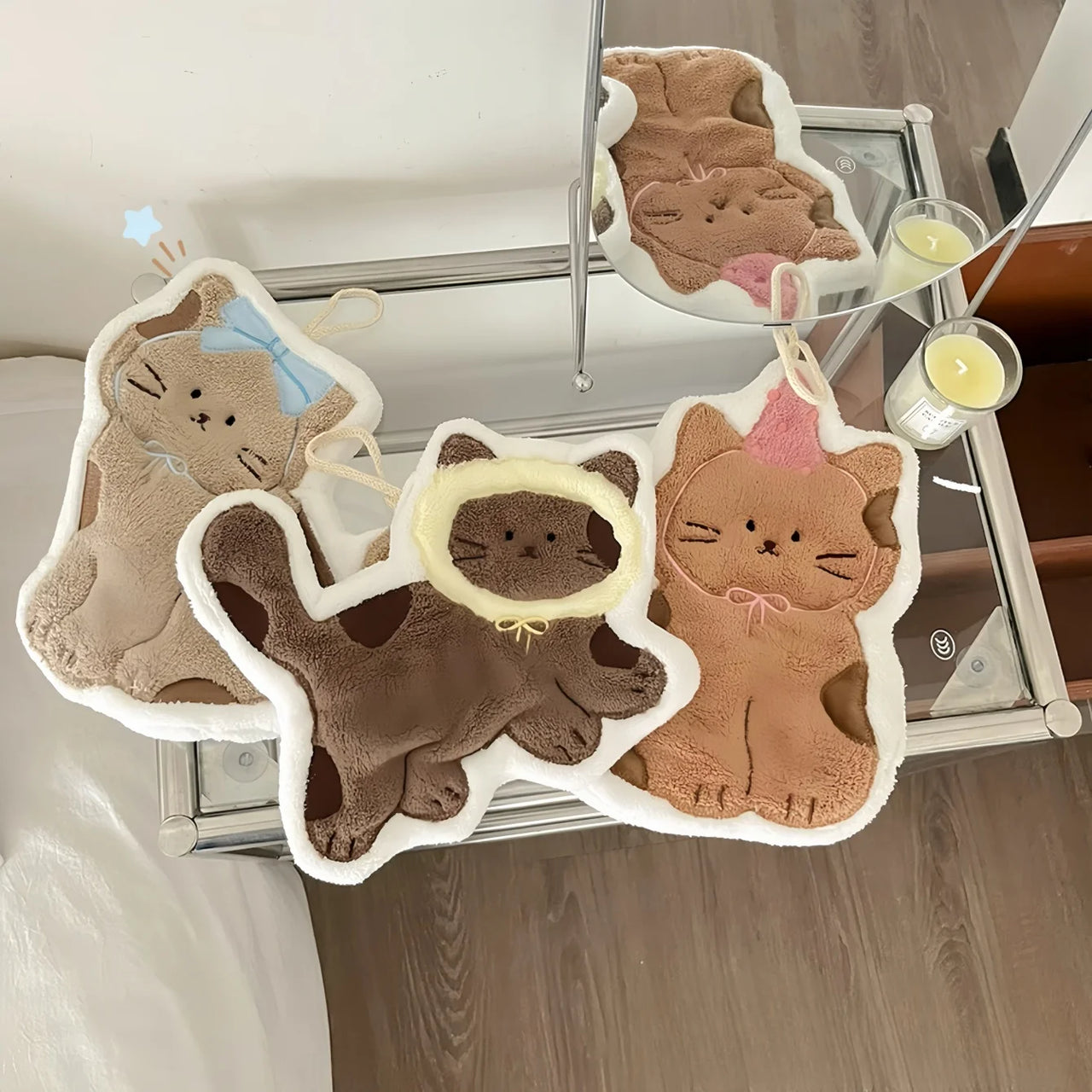 Three cat plushies with party hats are beside a KittyNook Cat Co. Soft Cat Paw Hand Towel on a glass table near candles.