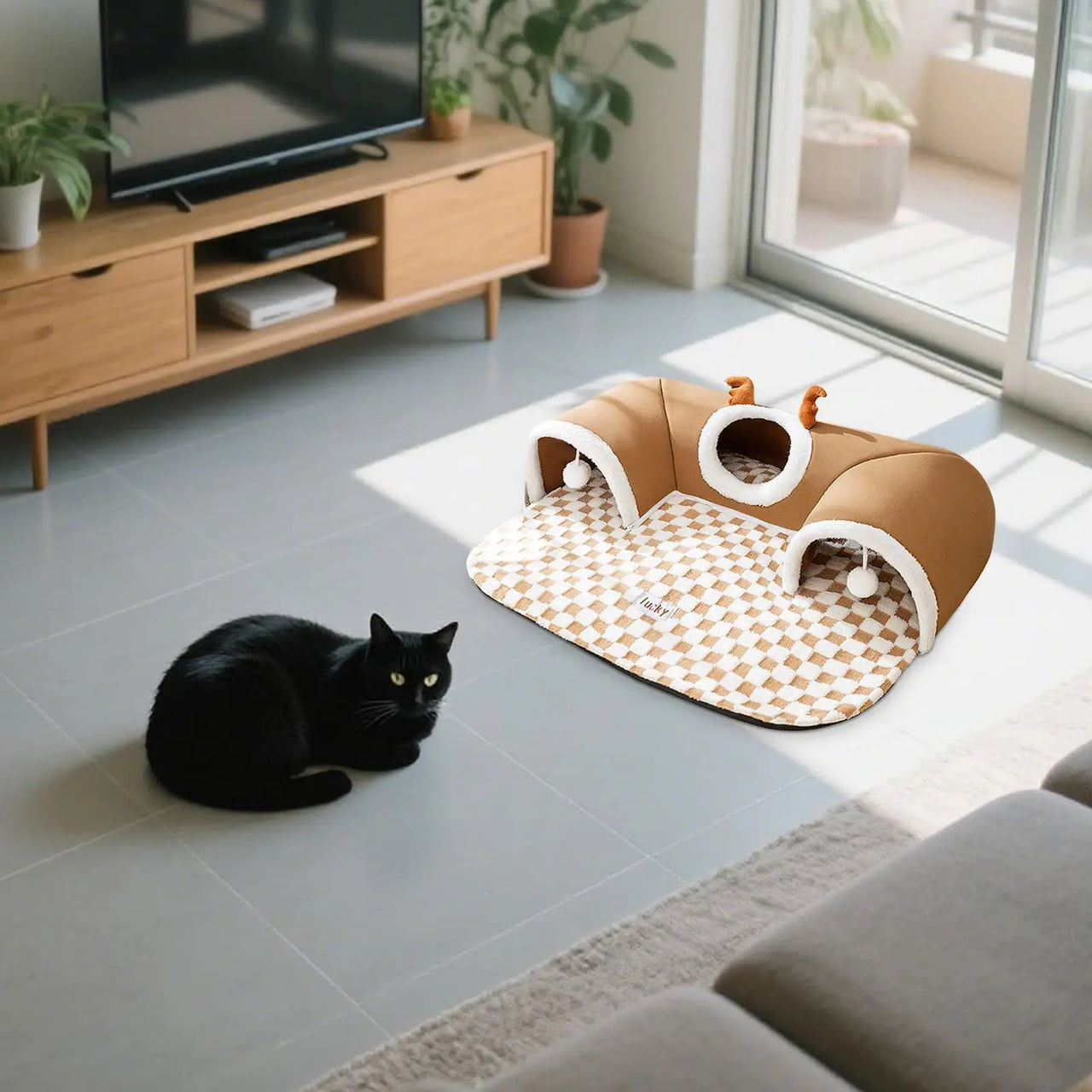 A black cat sits near a KittyNook Cat Co. Winter Warm Plush Tunnel Cat Bed in a bright living room.