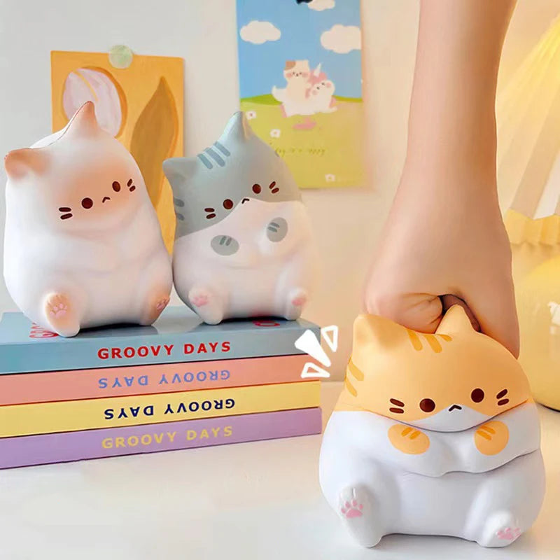 Three KittyNook Cat Co. Cat Squeeze Balls on books, with a hand squeezing one; fun pastel background decor.