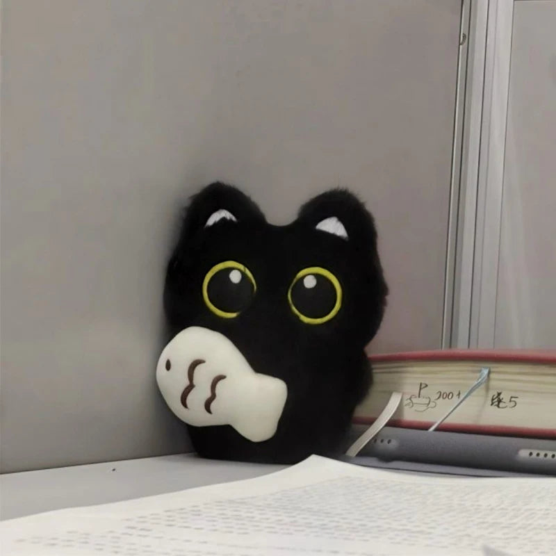 The KittyNook Cat Co. Black Cat Plush Doll Keychain Pendant with a fish and big yellow eyes leans on desk books.