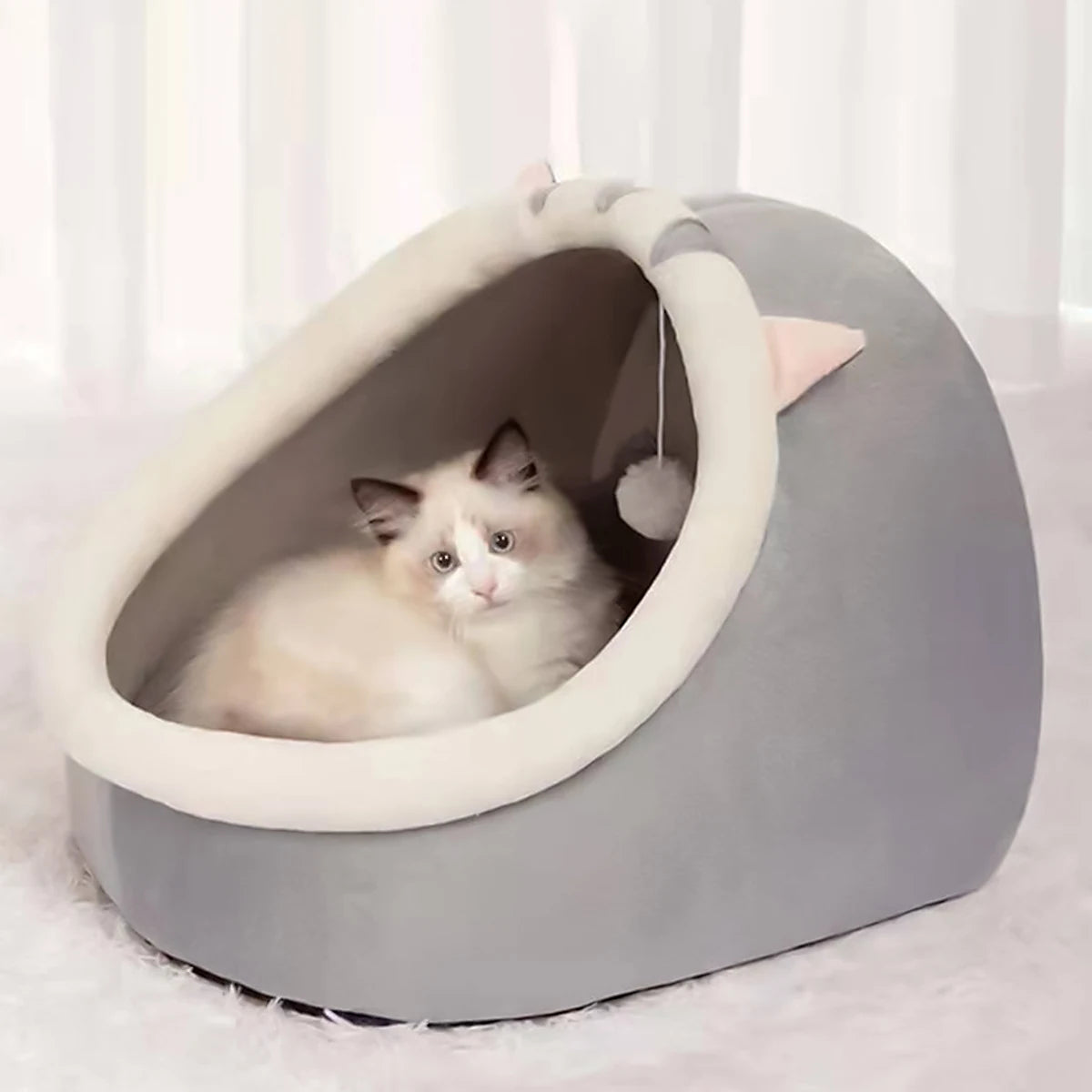 A white and gray cat lounges in the KittyNook Cat Co. Cute Comfortable Cat Nest Bed with pink ears and a hanging toy.