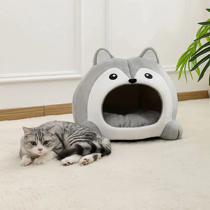 A gray tabby cat lies on a rug next to the KittyNook Cat Co. Fluffy Indoor Cat Cave Bed for Kittens in a bright room.