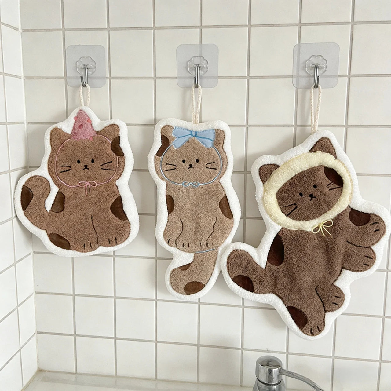 Three Soft Cat Paw Hand Towels by KittyNook Cat Co. hang on hooks above a white tiled sink.