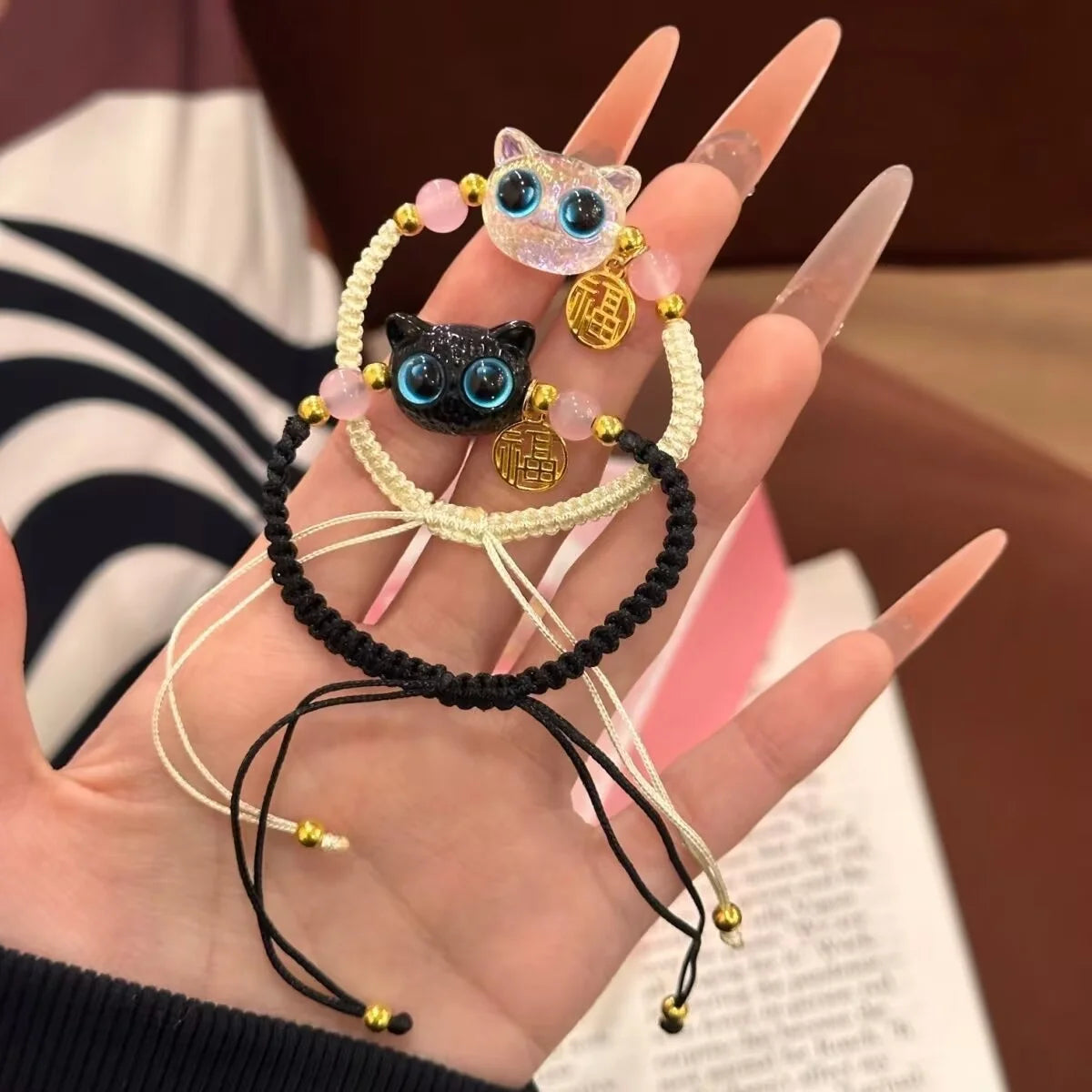 A hand holds two KittyNook Cat Co. Lucky Charm Bracelets—blue-eyed cat beads and gold charms, perfect for pet parents.
