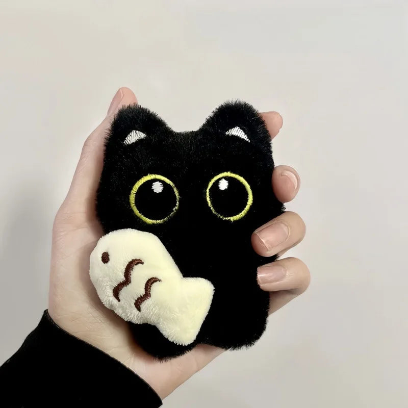 A hand holds the KittyNook Cat Co. Black Cat Plush Doll Keychain Pendant with a soft white fish in its mouth.