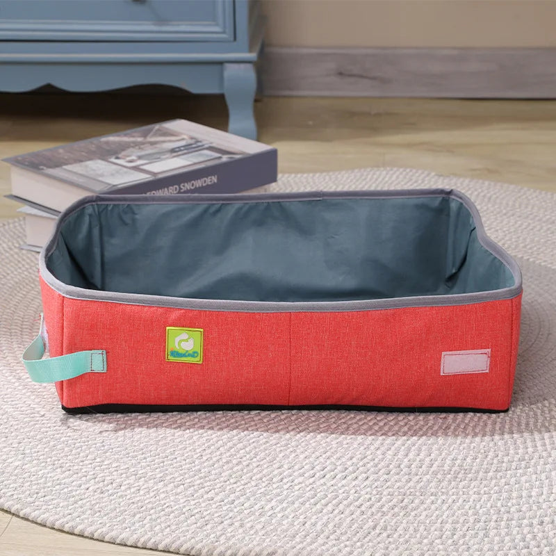 The KittyNook Cat Co. Portable Cat Litter Box in red and gray rests on a rug near books and a blue cabinet.