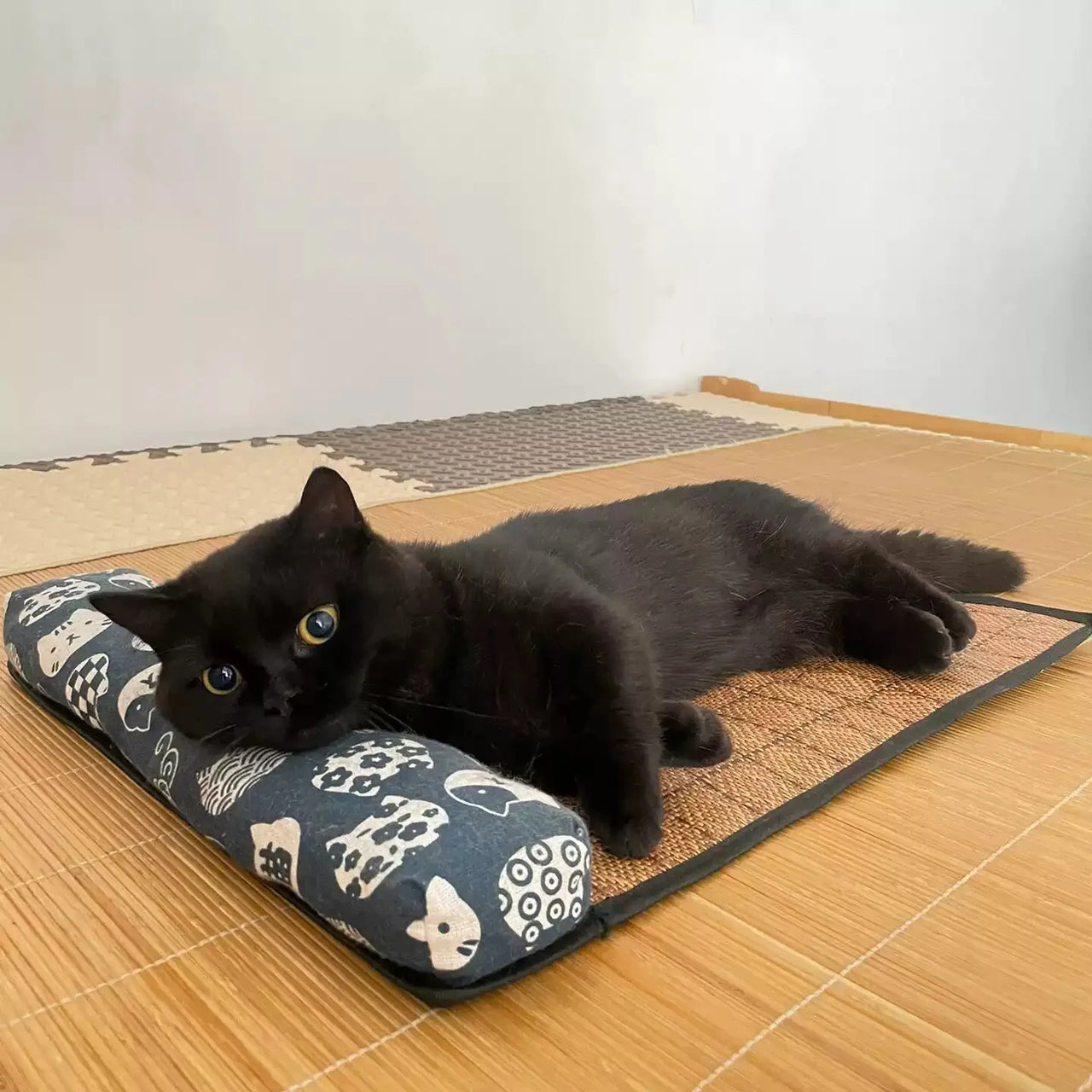 A black cat lounges on the KittyNook Cat Co. Natural Rattan Cat Sleeping Mat, gazing up with wide eyes.