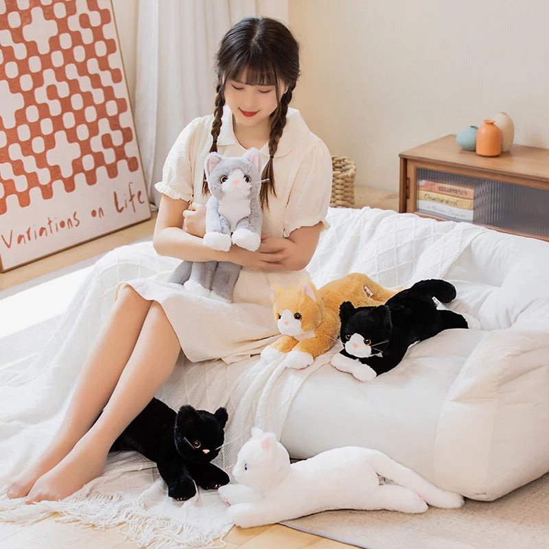 A woman in white sits on a bed, hugging a KittyNook Cat Co. Snuggle Soft Cat Plush Doll, surrounded by others.