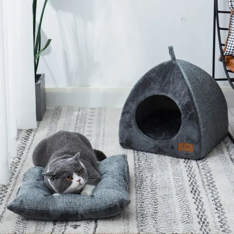 A gray cat lounges on a gray cushion near the KittyNook Cat Co. Cozy Triangle Cat Cave Bed in a modern room.