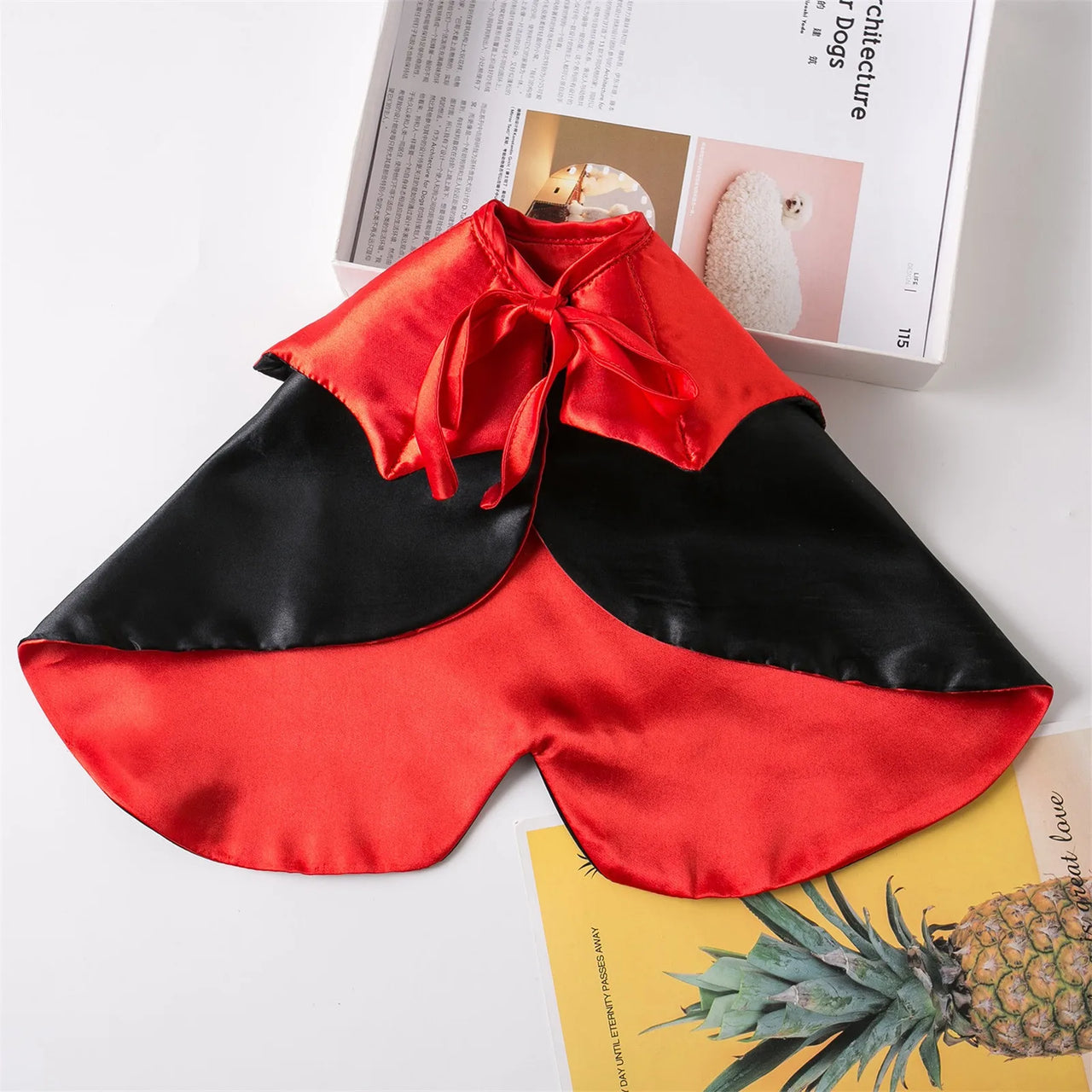 KittyNook Cat Co. Cute Halloween Cat Costumes: red and black satin cape with ribbon tie, near magazines and pineapple print.