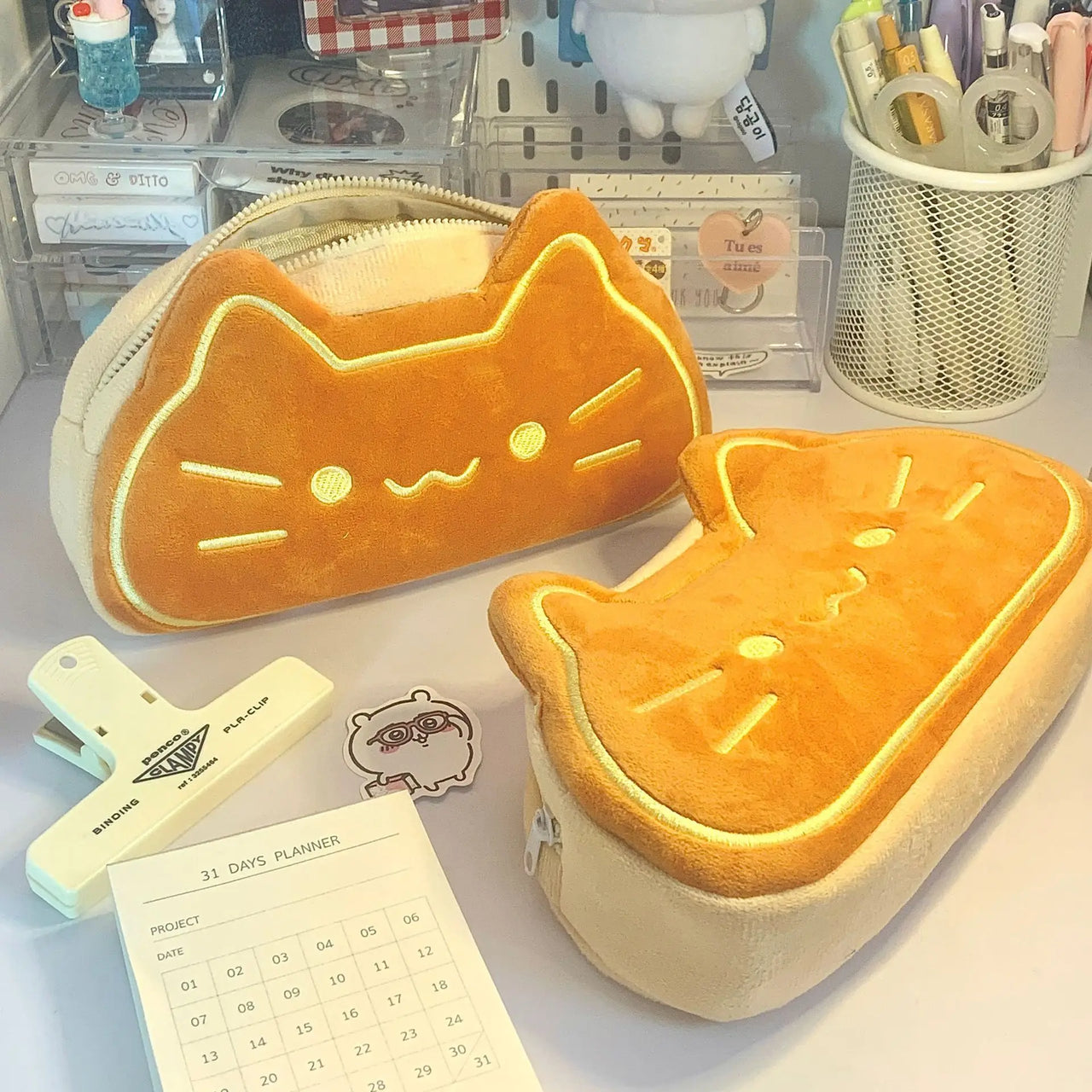 Two cute pencil cases, including KittyNook Cat Co.'s Caramel Cat Plush Pencil Case, sit on a desk with a 31-day planner.