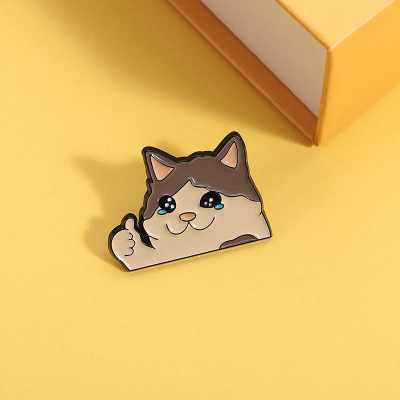 KittyNook Cat Co.’s Kawaii Thumbs Up Crying Cat Enamel Pin on yellow beside an orange and white box.