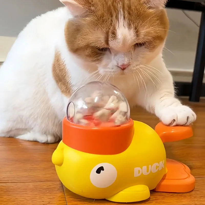 Orange and white cat taps a yellow KittyNook Cat Co. Non-Toxic Pet IQ Food Leak Tumbler on a wooden floor.