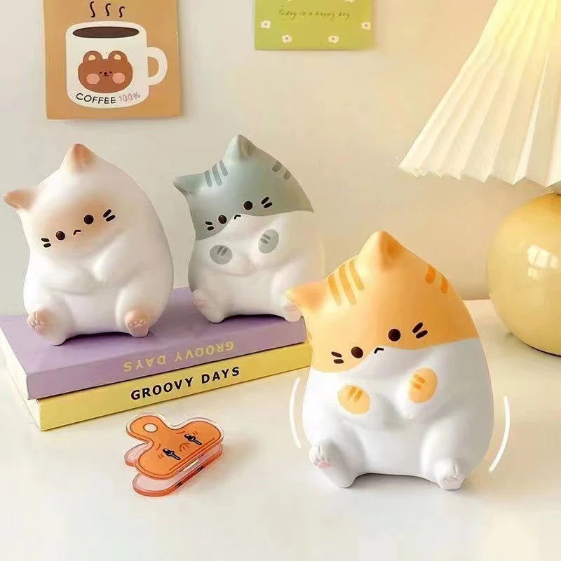 Three chubby cat figurines sit on books by a lamp and a KittyNook Cat Co. Cat Squeeze Ball for stress relief.