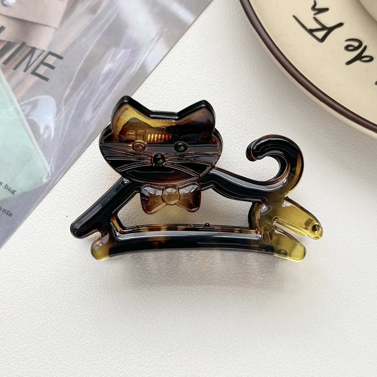 The KittyNook Cat Co. Cute Cat Hair Claw with a bowtie is on a white surface next to a plate and magazine.