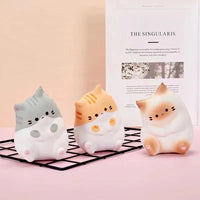 Thumbnail for Three KittyNook Cat Co. Cat Squeeze Balls in gray, orange, and brown sit on a pink surface with a magazine nearby.