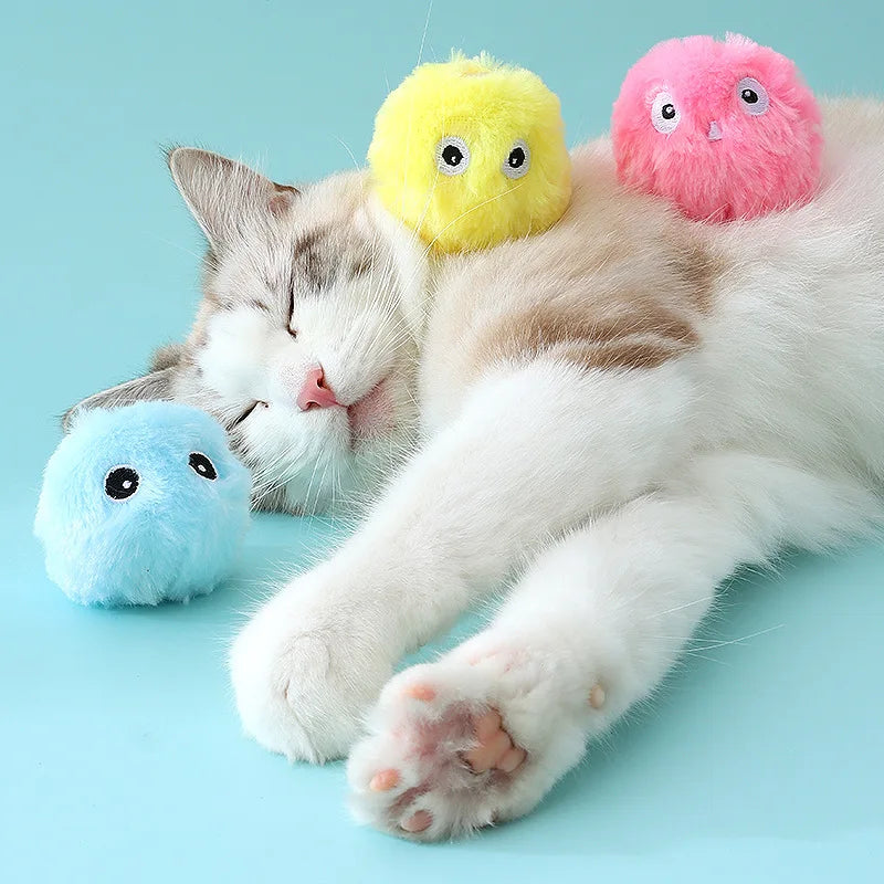 A cat sleeps on its side with plush toys and a KittyNook Cat Co. Interactive Cat Smart Ball Toy on a light blue background.