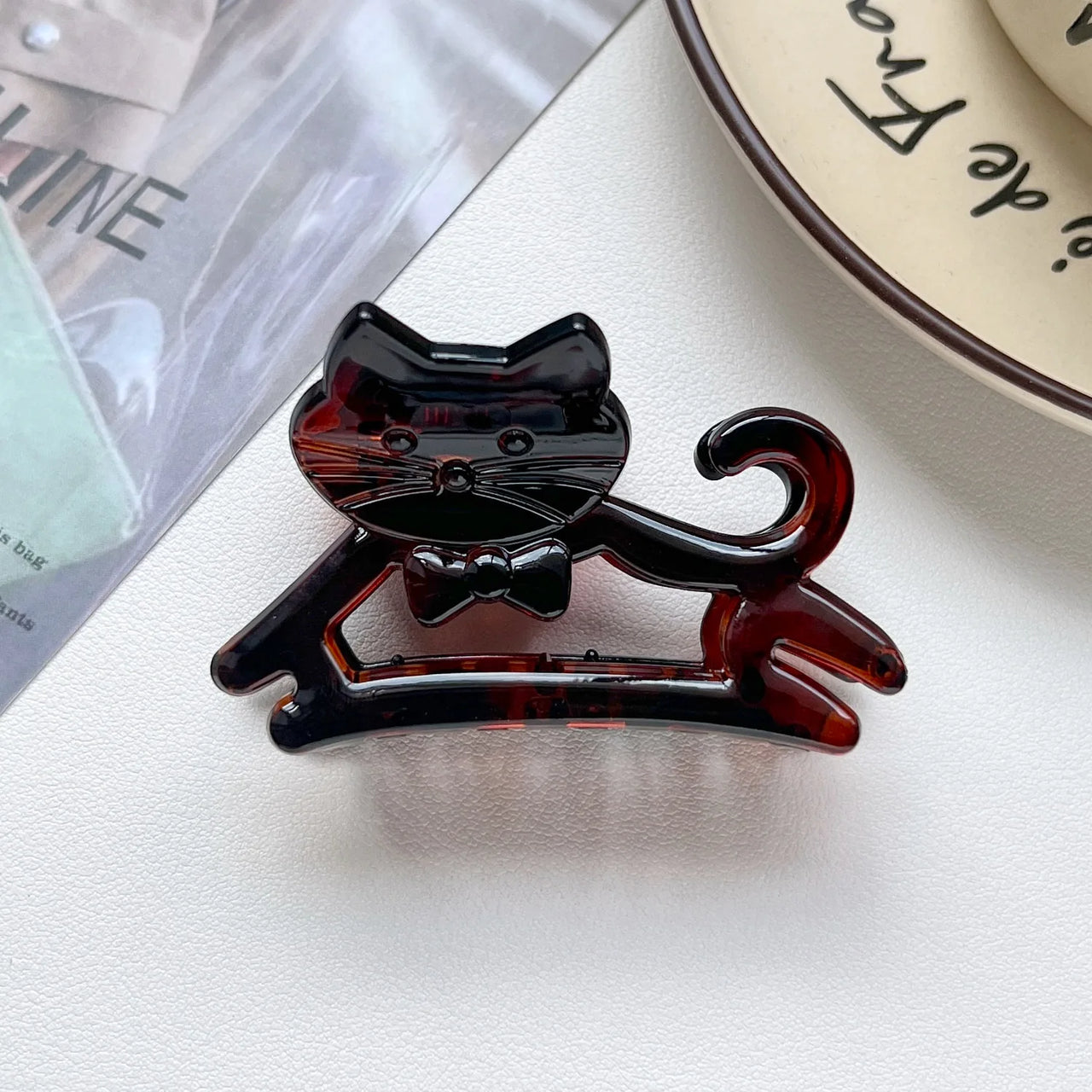 The KittyNook Cat Co. Cute Cat Hair Claw with a black bow tie sits on a white surface by a plate and magazine.