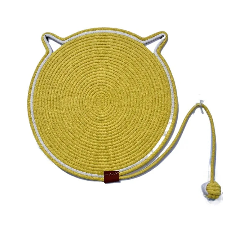 The Tails Cat Scratching Mat by KittyNook Cat Co. is a round yellow cat face trivet with ears and a long cord ending in a ball.