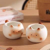 Thumbnail for Two round Siamese Cat Coffee Cups by KittyNook Cat Co., with cute faces and ears, sit on a wooden table near a book.