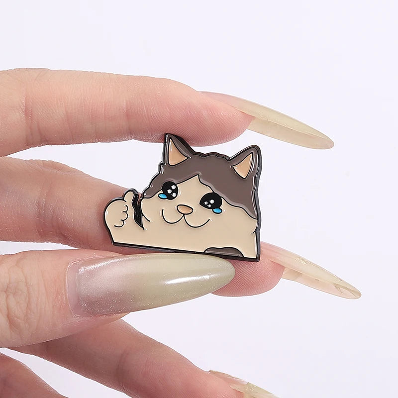 A hand with long nails holds KittyNook Cat Co.’s Kawaii Thumbs Up Crying Cat Enamel Pin—a cute smiling kitty accessory.