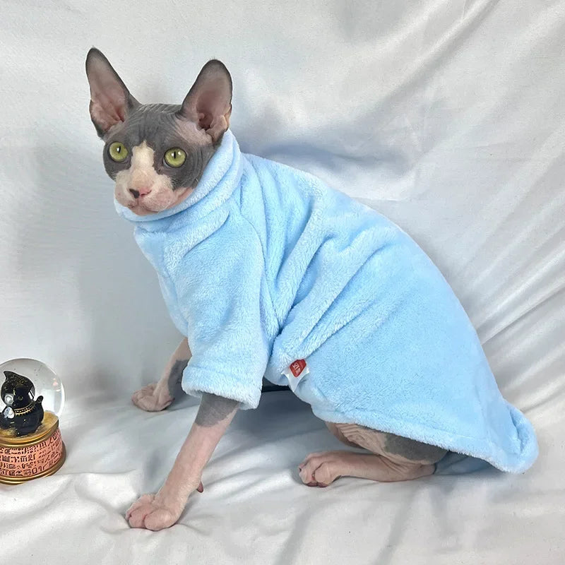 A hairless kitten in a Warm Winter Hoodie by KittyNook Cat Co. sits on a white surface beside a cat snow globe.