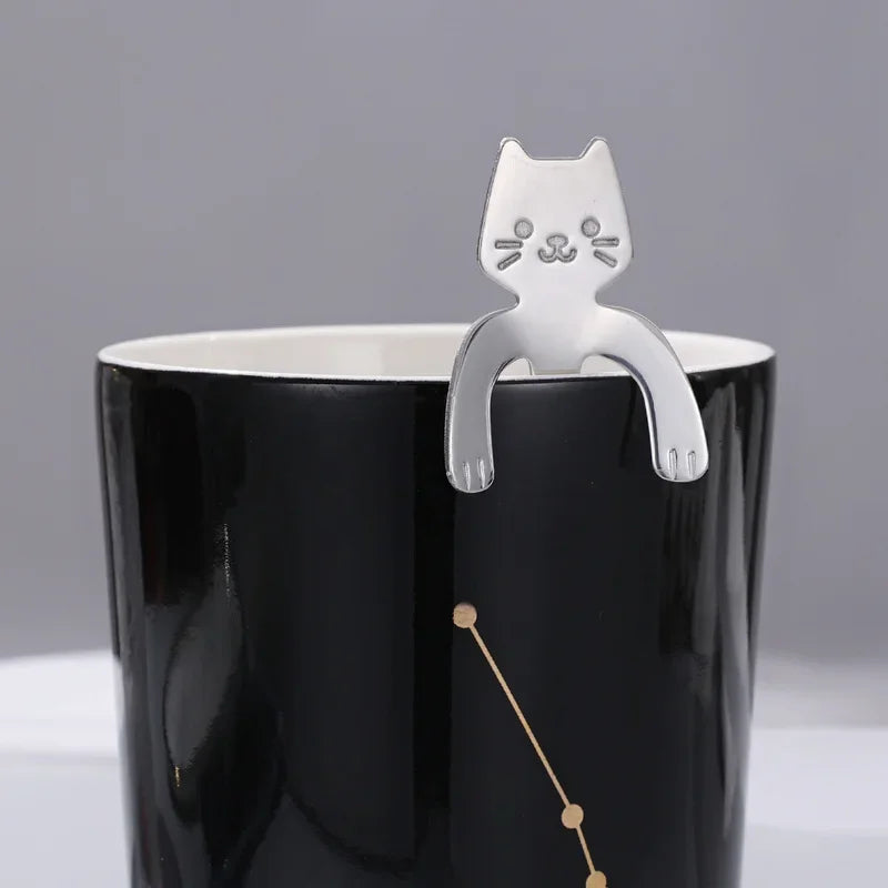 The KittyNook Cat Co. Cat Cling Stainless Steel Mug Spoon hangs on a black mug with a gold constellation design.