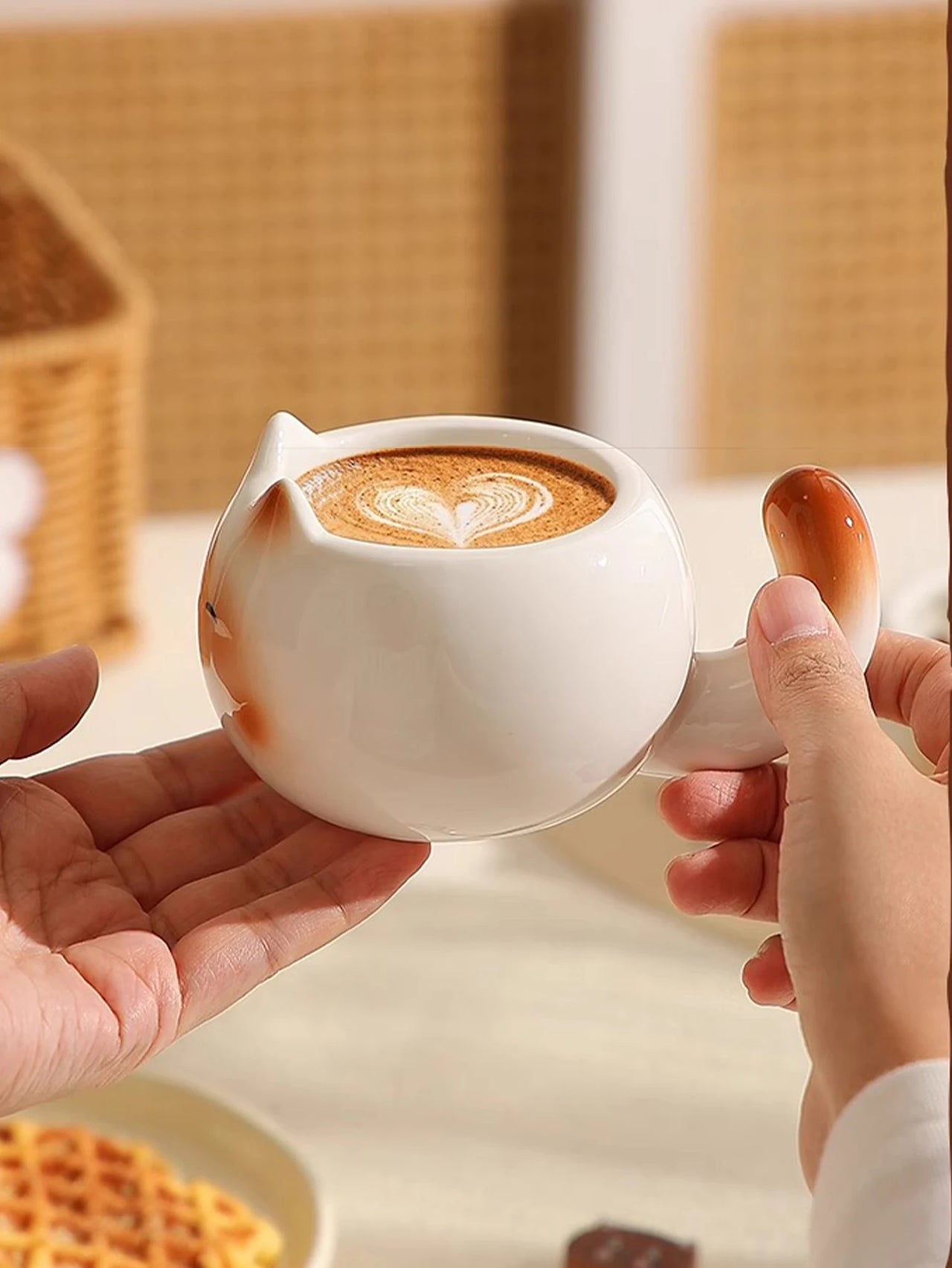 Two hands hold a KittyNook Cat Co. Siamese Cat Coffee Cup with heart latte art, waffles in the background.