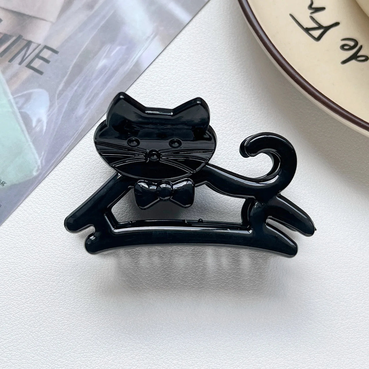 The KittyNook Cat Co. Cute Cat Hair Claw with bow tie is a chic K-style accessory, shown near a plate and magazine.
