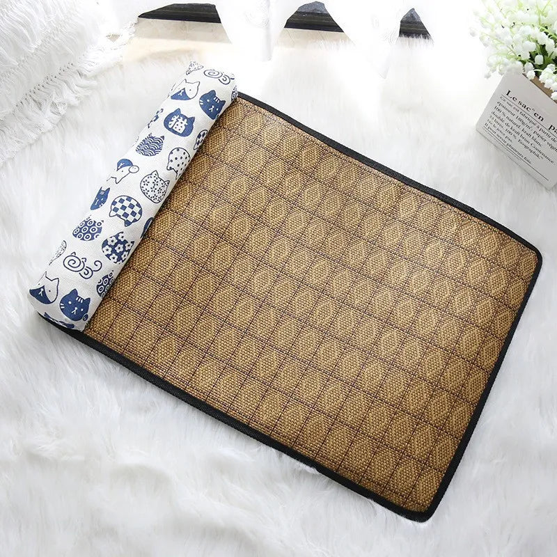 The KittyNook Cat Co. Natural Rattan Cat Sleeping Mat with a blue and white pillow sits on a fluffy surface by the window.