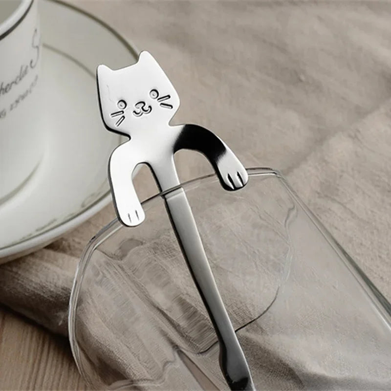 The KittyNook Cat Co. Cat Cling Stainless Steel Mug Spoon hangs on a glass, beside a white cup on beige cloth.