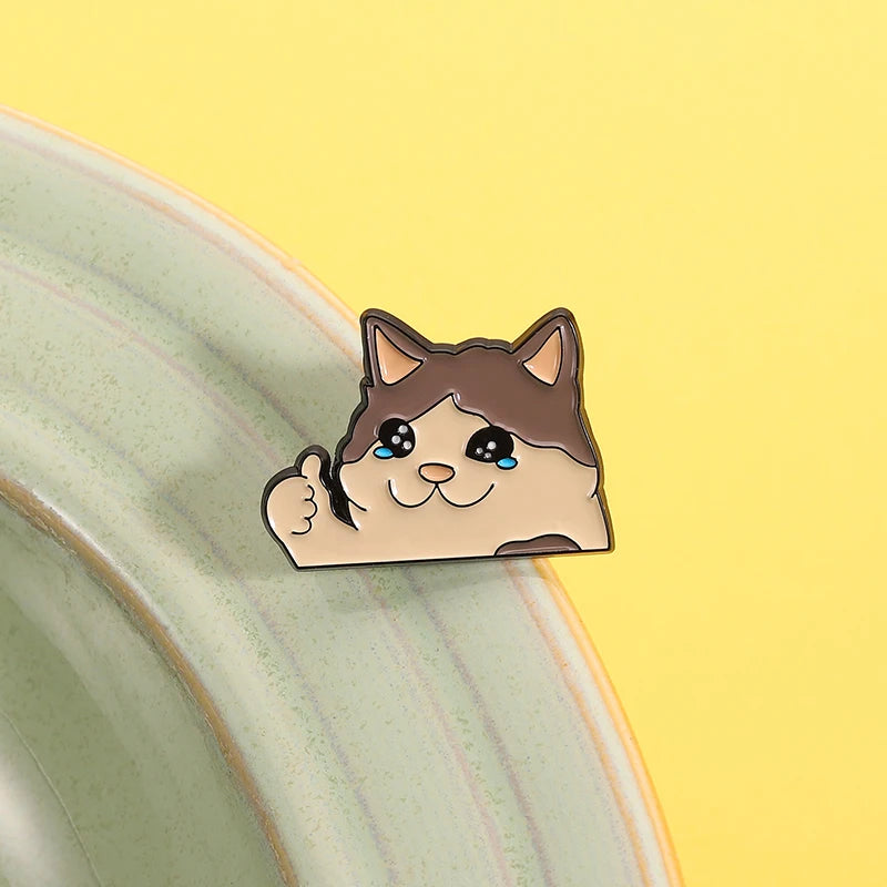Kawaii Thumbs Up Crying Cat Enamel Pin by KittyNook Cat Co. sits on a green bowl, yellow background cheers it up.