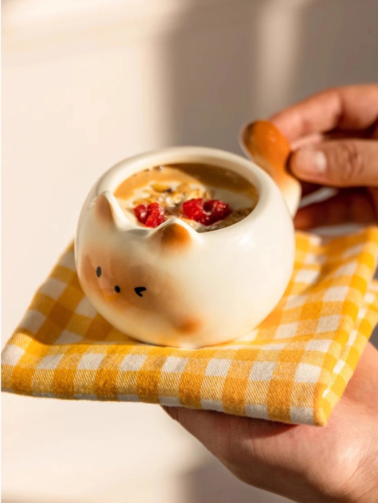 A hand holds the KittyNook Cat Co. Siamese Cat Coffee Cup with oatmeal and berries on a yellow checkered cloth.