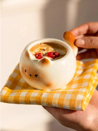 Thumbnail for A hand holds the KittyNook Cat Co. Siamese Cat Coffee Cup with oatmeal and berries on a yellow checkered cloth.