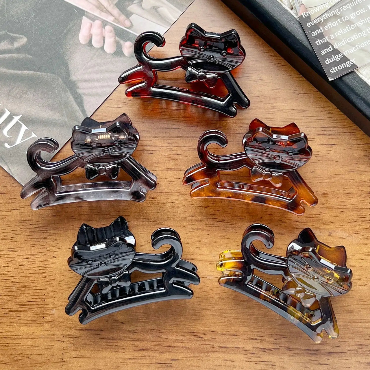 Six KittyNook Cat Co. Cute Cat Hair Claws in assorted colors rest on a wooden surface by a magazine and book.